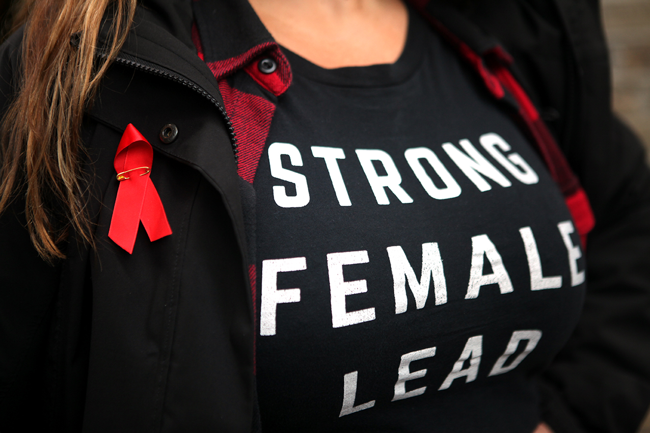Chest of woman with red ribbon (AIDS symbol) pinned to clothes. T shirt reads 'strong female lead'